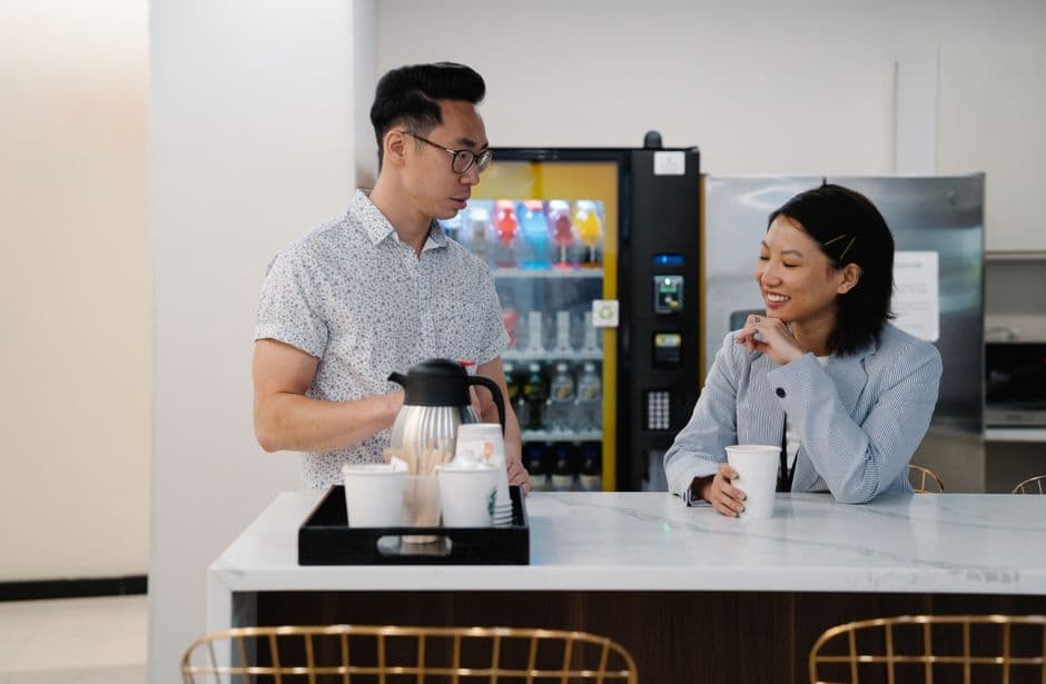 A casual interaction between colleagues at an office coffee station with a vending machine.