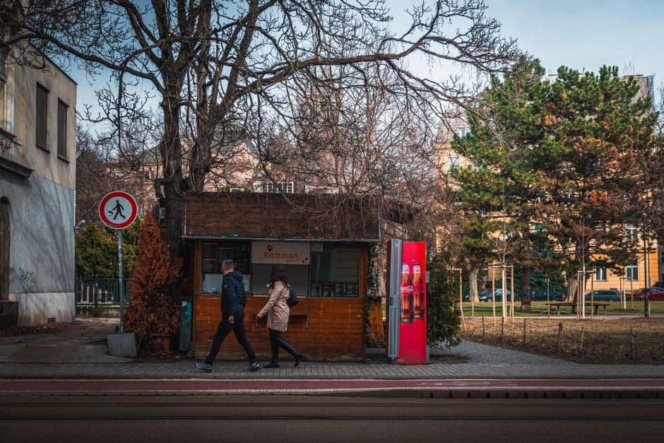 City street scene featuring pedestrians walking past a kiosk with bare trees in winter.