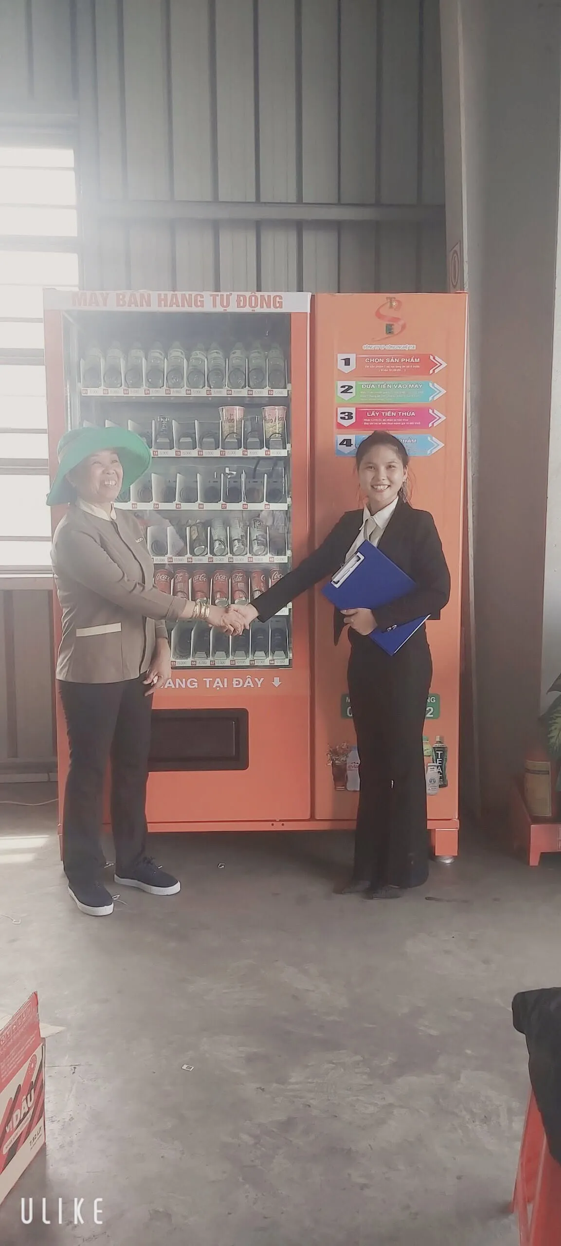 Hai đại diện bắt tay trước máy bán hàng tự động màu cam trong khu vực nhà xưởng, đánh dấu bàn giao thành công
