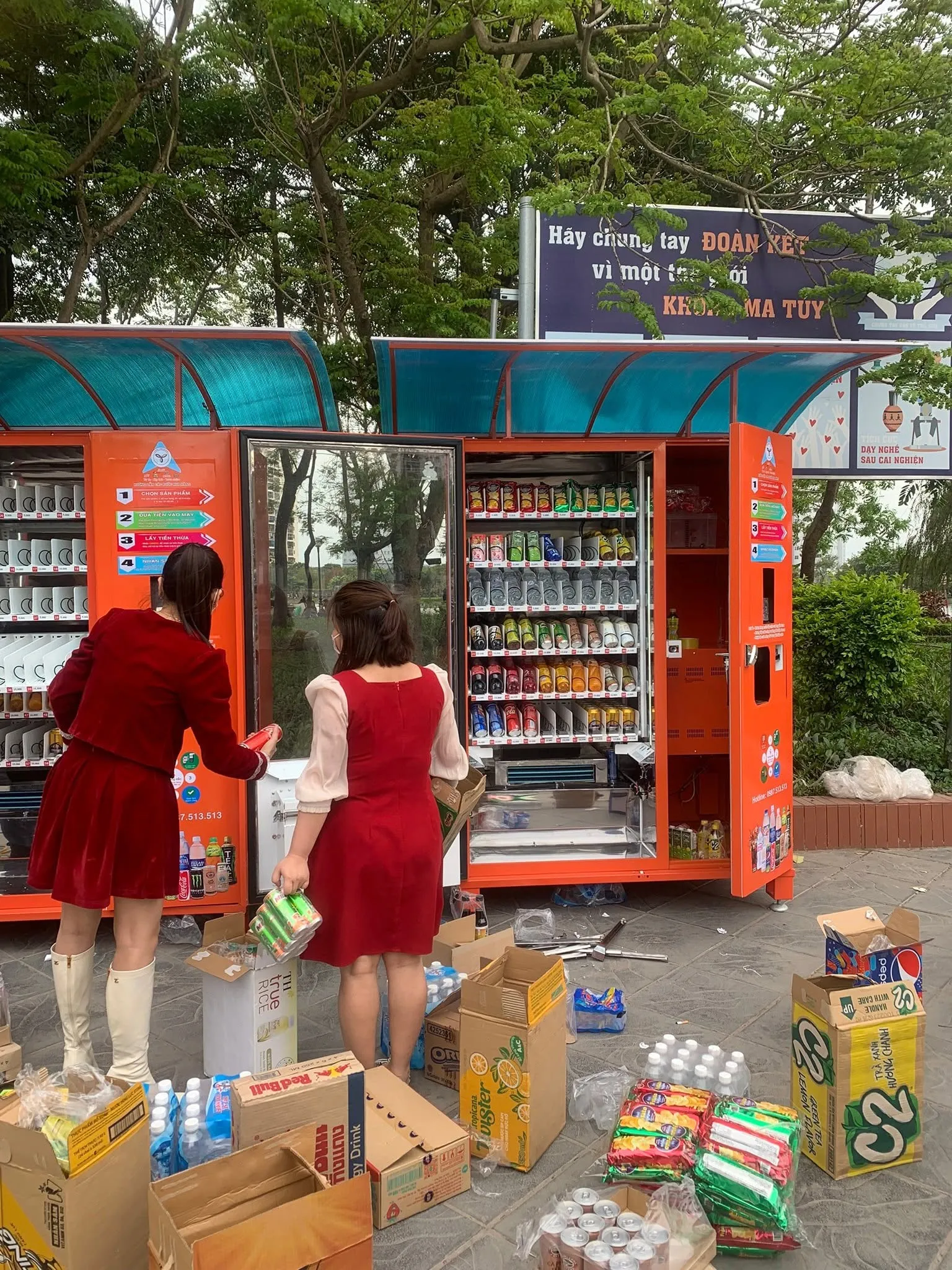 Hai nhân viên châm hàng cho cụm máy đôi bán nước và đồ ăn nhẹ tại công viên, nhiều thùng hàng để xung quanh