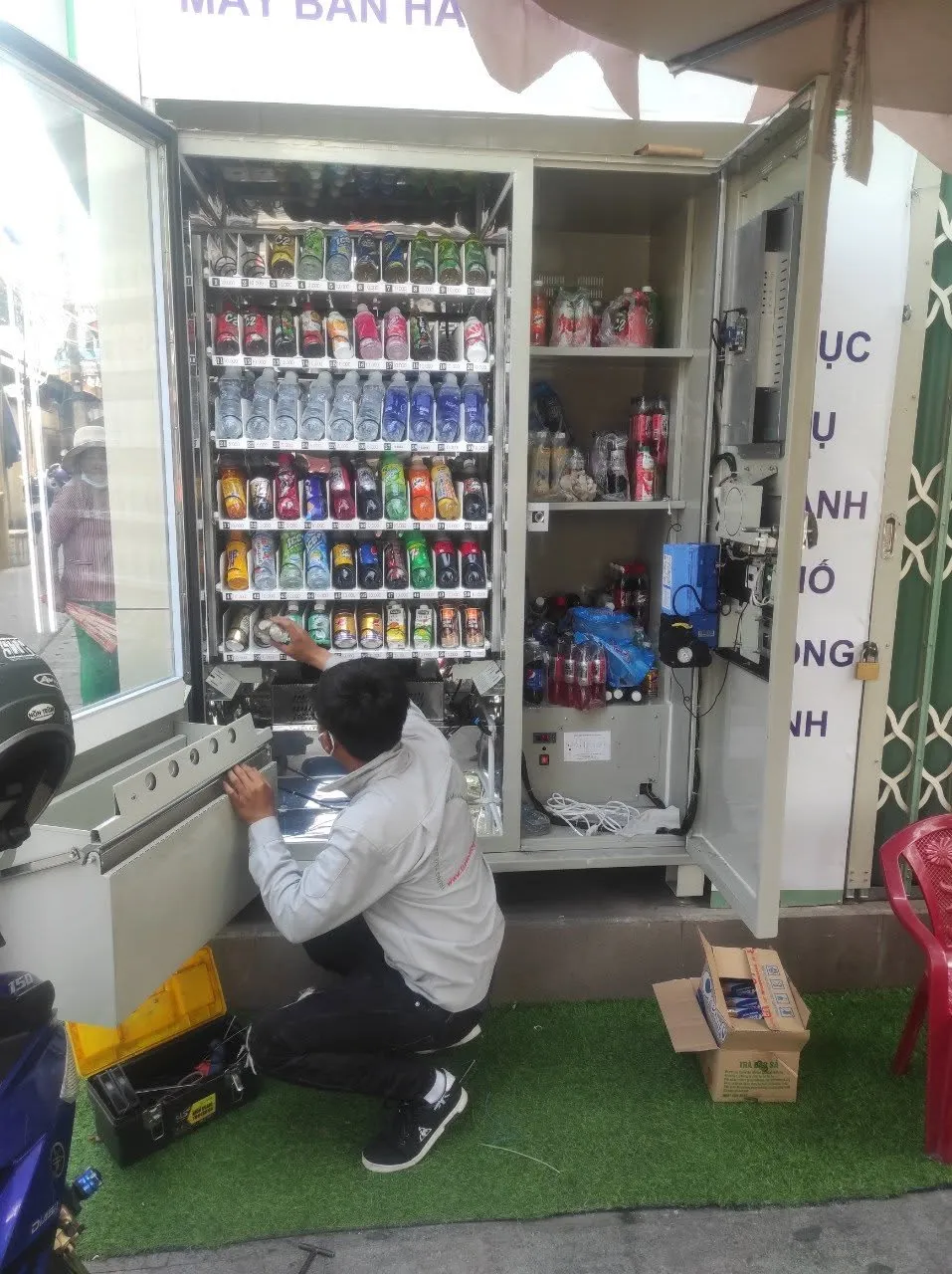 Kỹ thuật viên quỳ châm hàng cho máy bán hàng tự động đang mở cửa, bên trong đầy nước ngọt và snack