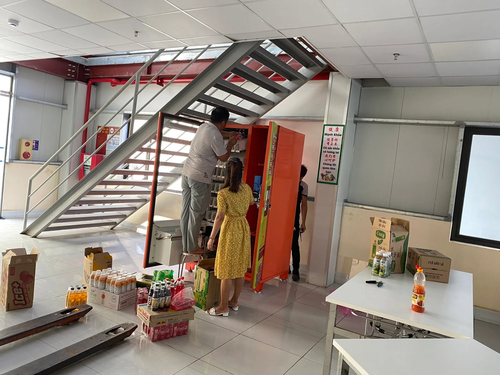 Lắp đặt và nạp hàng cho máy bán hàng tự động đặt dưới gầm cầu thang trong tòa nhà, hàng hóa để sẵn bên cạnh