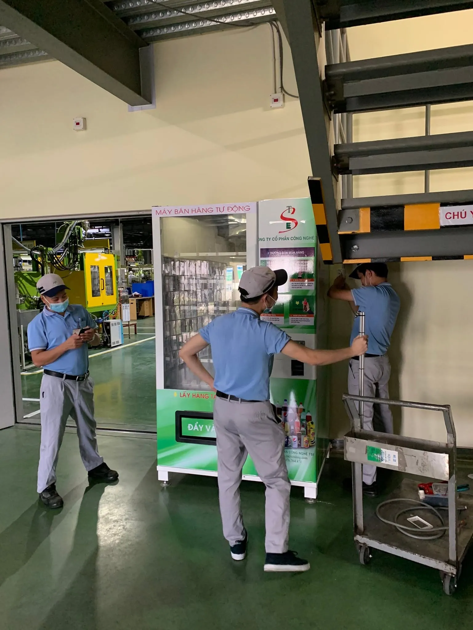 Đội kỹ thuật áo xanh đưa máy bán hàng tự động vào vị trí lắp đặt bên trong nhà máy sản xuất