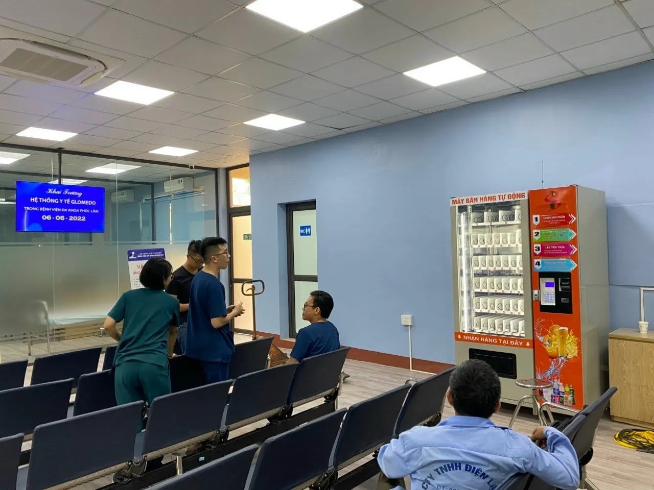 Máy bán hàng tự động màu đỏ đặt tại phòng chờ bệnh viện đa khoa, có ghế chờ và màn hình thông báo