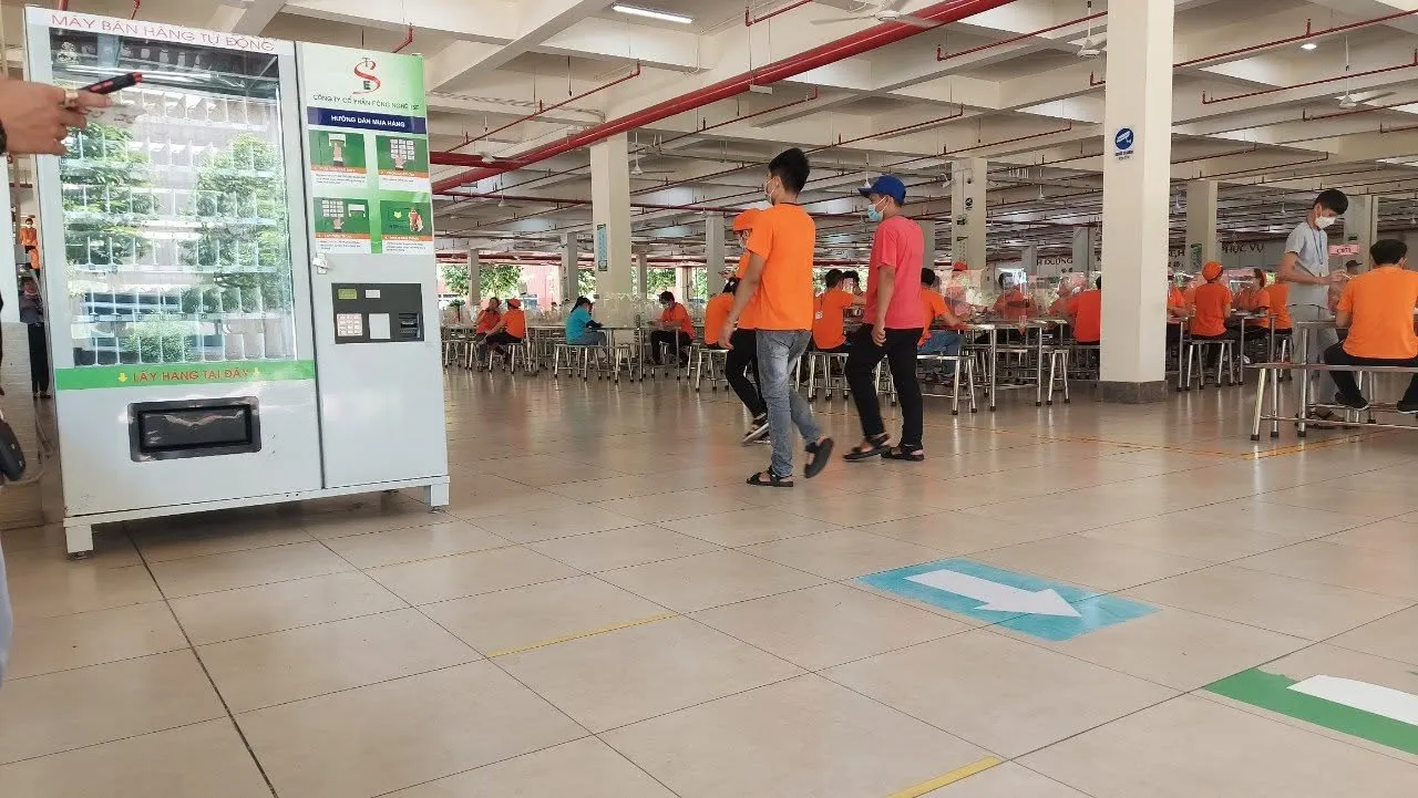 Máy bán hàng tự động đặt trong căng tin nhà máy, công nhân mặc áo cam đứng xung quanh