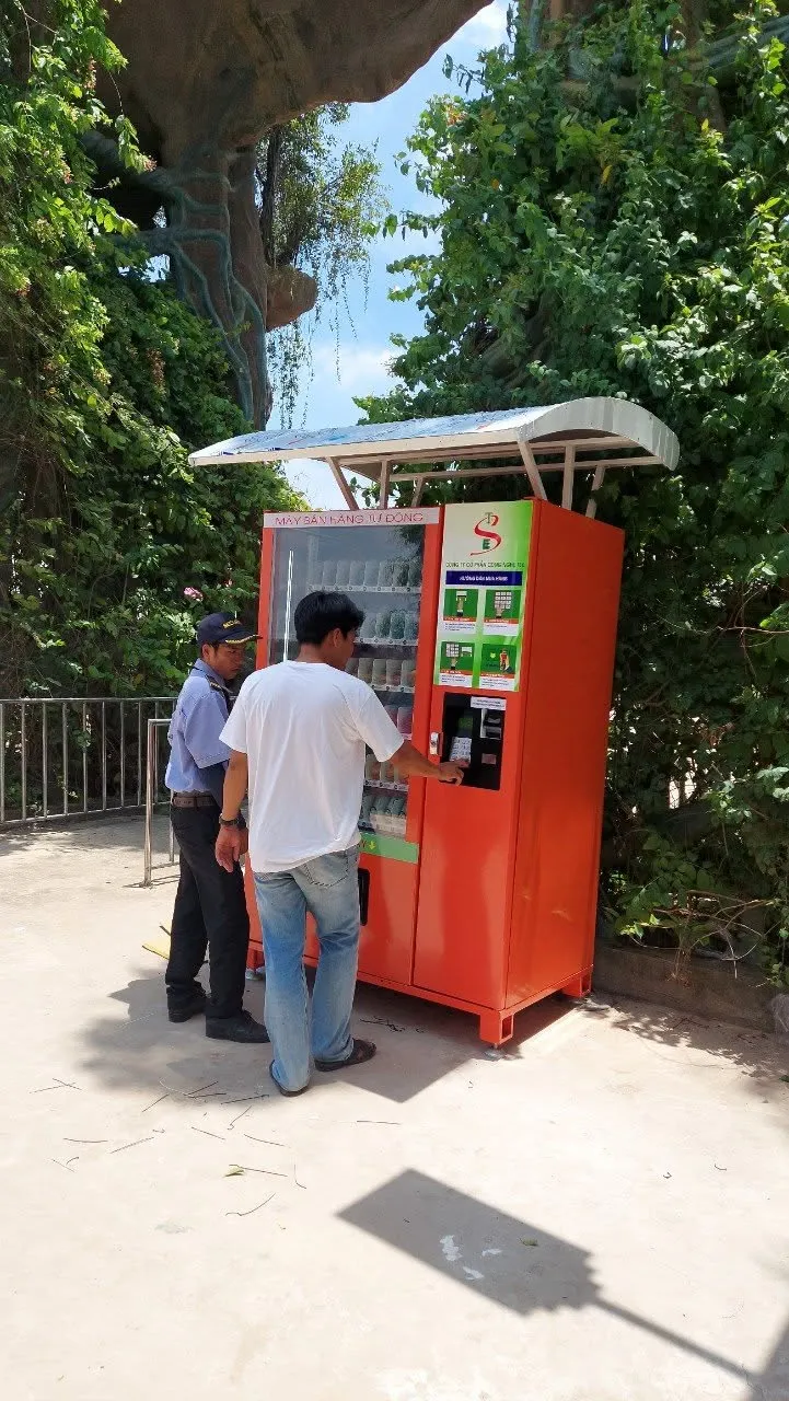 Máy bán hàng tự động màu cam có mái che gắn tấm năng lượng mặt trời đặt trong công viên, khách và bảo vệ đứng cạnh