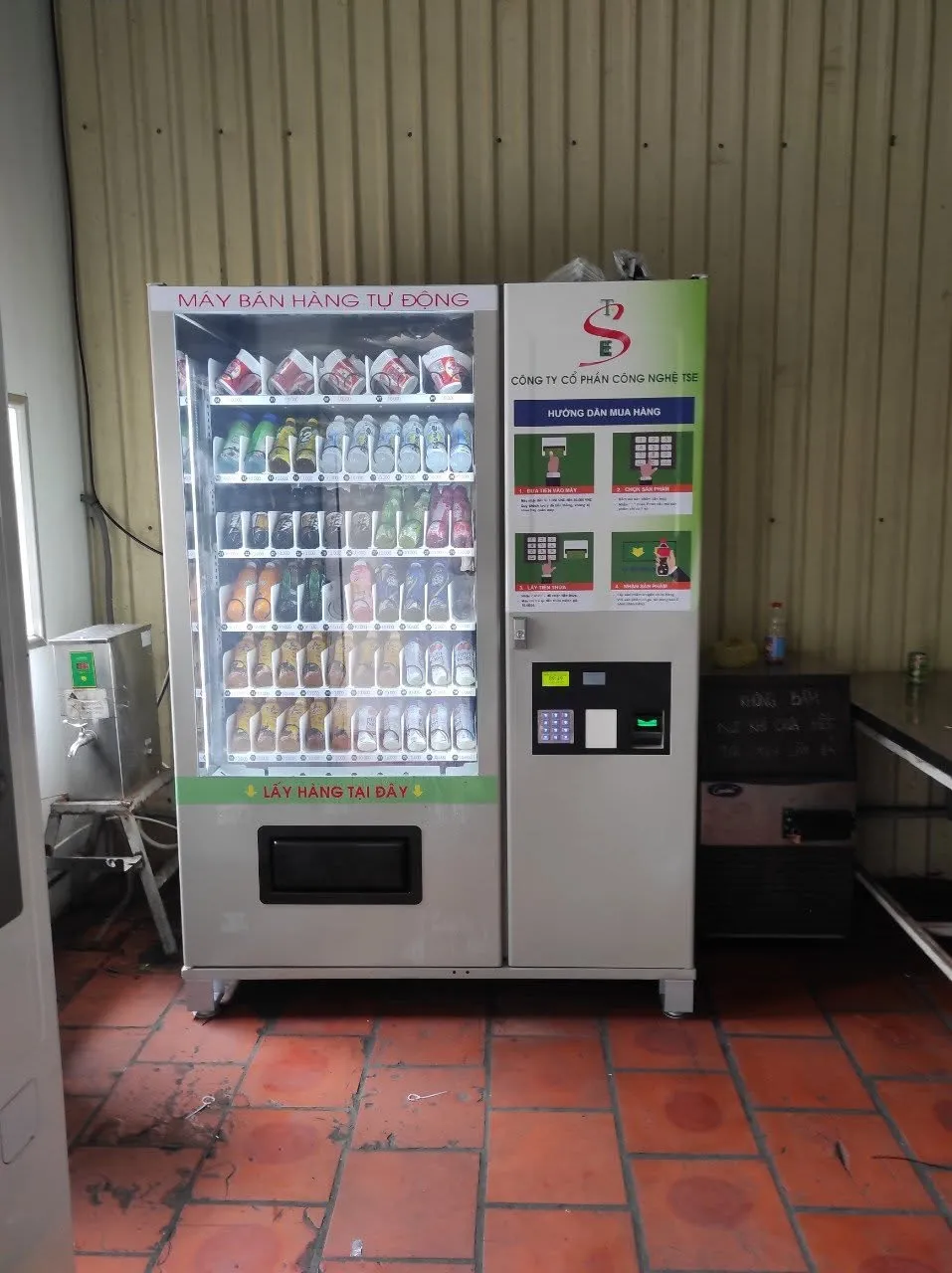 Máy bán hàng tự động TSE đặt trong nhà trên nền gạch đỏ, bán nước và đồ ăn nhẹ, có bảng hướng dẫn