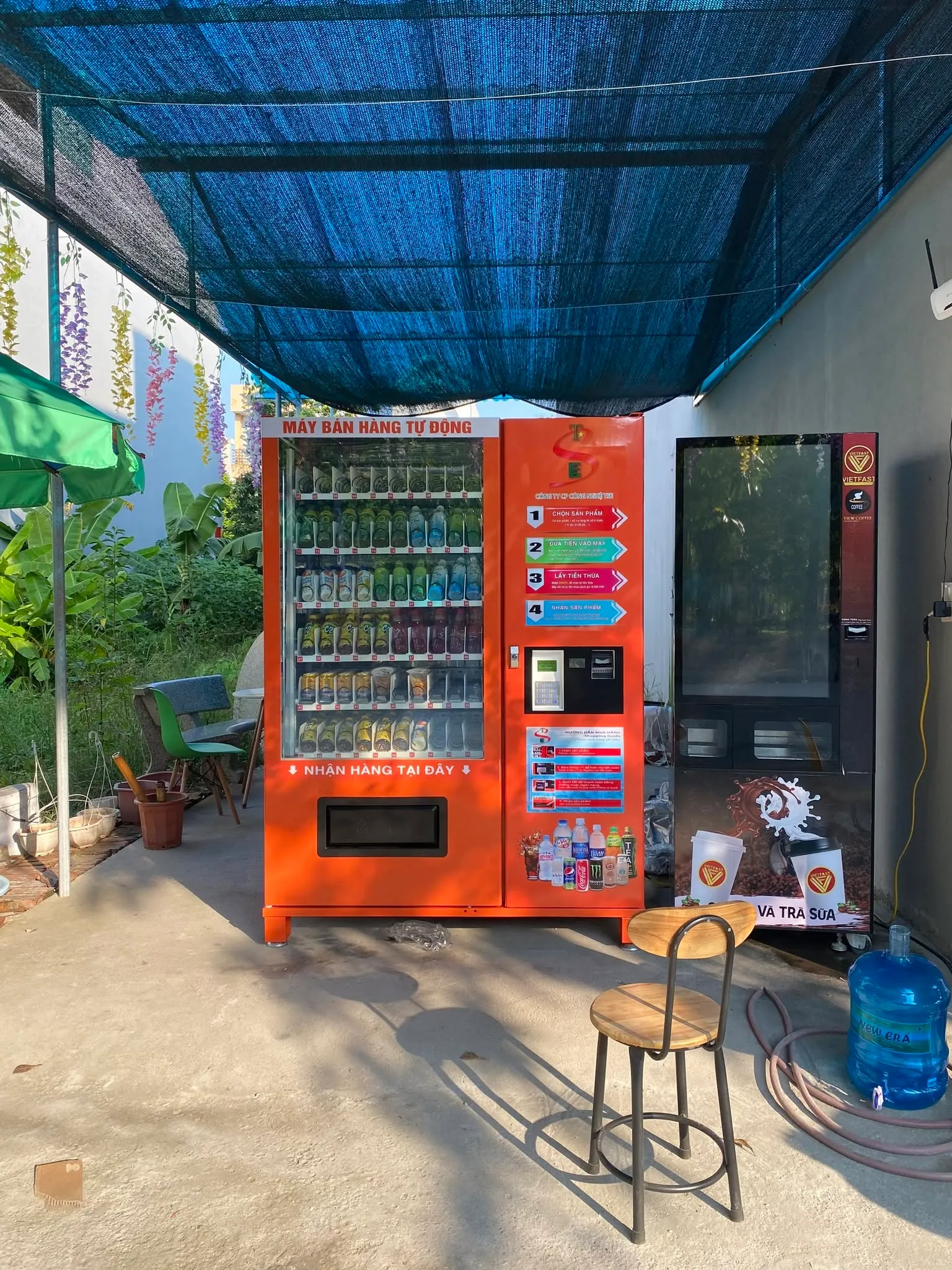 Máy bán hàng tự động màu cam đặt tại sân vườn quán cà phê dưới ô che nắng, có bàn ghế xung quanh