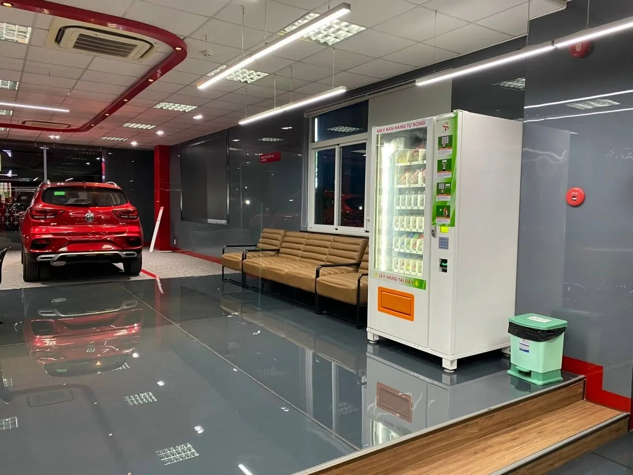 Máy bán hàng tự động đặt tại khu vực chờ của showroom ô tô, phía sau là xe trưng bày màu đỏ