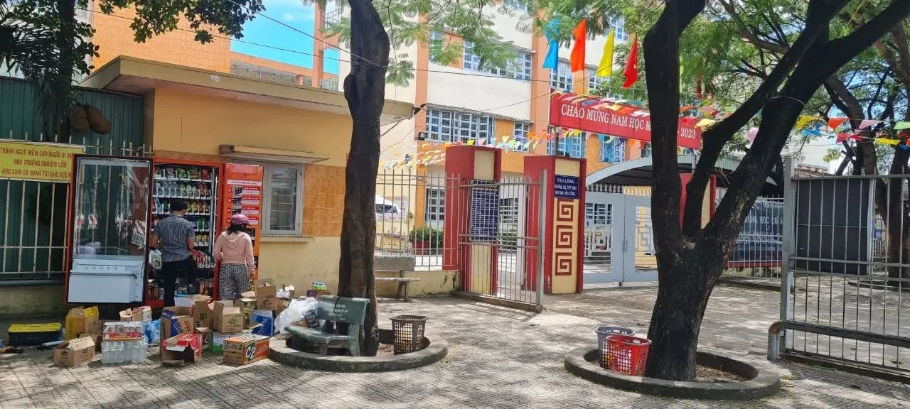 Máy bán hàng tự động đặt gần cổng trường học có băng rôn chào mừng năm học mới, cạnh quầy tạp hóa