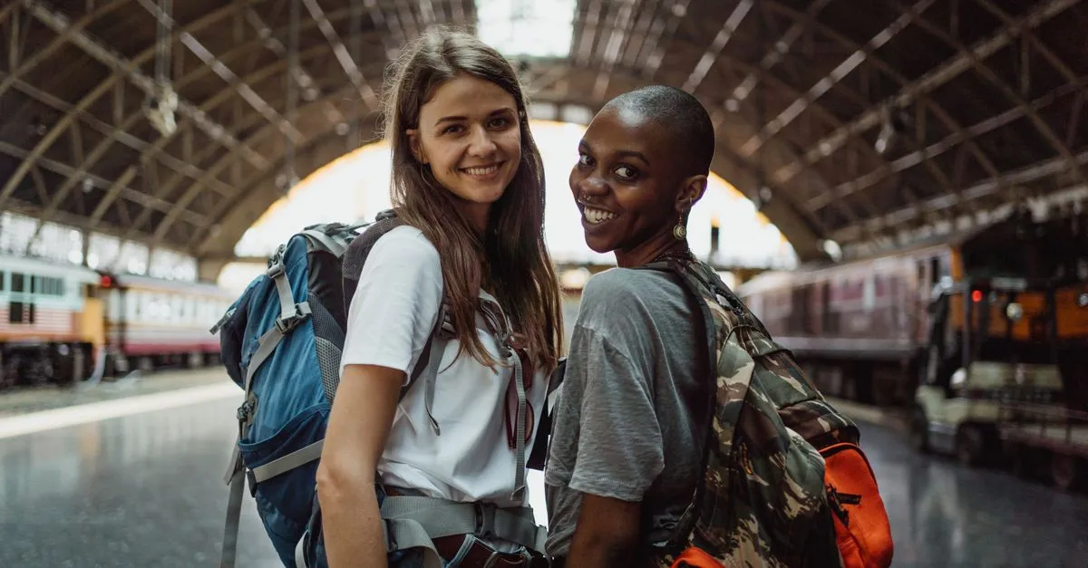 Two women with backpacks smiling at a train station, ready for their travel adventure.
